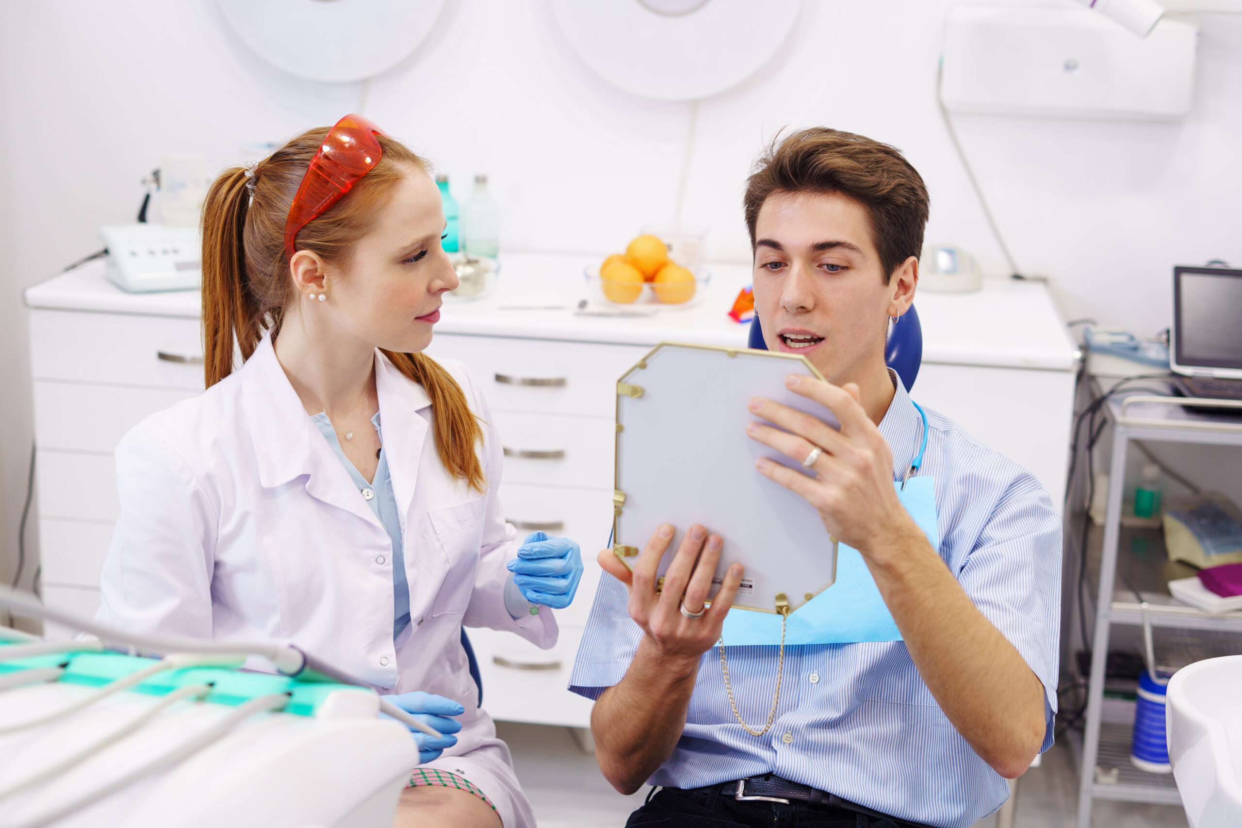 Male patient looking at teeth reflection in mirror while sitting near redhead female doctor in modern dentist office